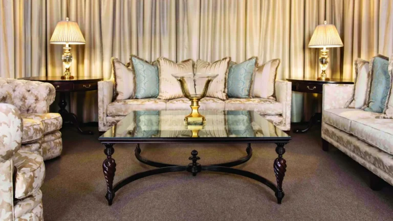 Elegant living room furniture set featuring a dark wood glass-top coffee table with spiral legs, surrounded by beige upholstered sofas with light blue throw pillows, floral armchairs, and matching table lamps against floor-to-ceiling curtains.